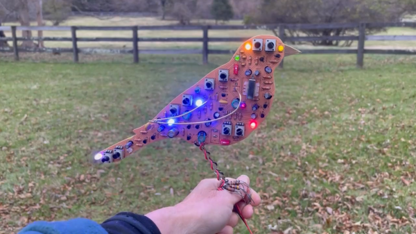 A bird-shaped yellow PCB with legs wound out of wire, perched on its creator's arm. The bird has a lot of through-hole components on it, as well as an assortment of different-colored LEDs.
