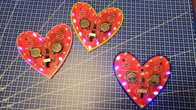Three pendants that the article is describing, on a drafting mat.They're heart-shaped red PCBs with LEDs all around its perimeter, two CR2032-like batteries in its center.