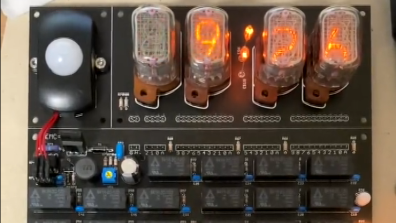 A black PCB with four numeric Nixie tubes on the top, showing 9:26. Under them, a group of black relays is located.