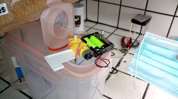 Mask DIY sanitization device on the left, mask used as an example on the right. The device is a Tupperware-like plastic container, on top, a small motor plus battery device with an alligator clip attached to the motor. Mask is inserted into the container through the opening on top, hooked to the motor, and the motor then spins the mask inside the container where hydrogen peroxide vapor is being misted.