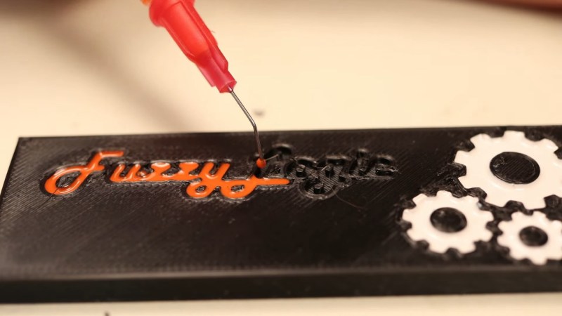 Syringe with diluted nail polish used to fill into cursive "FuzzyLogic" letters extruded into a surface of a 3D-printed block of plastic, as a demonstration.
