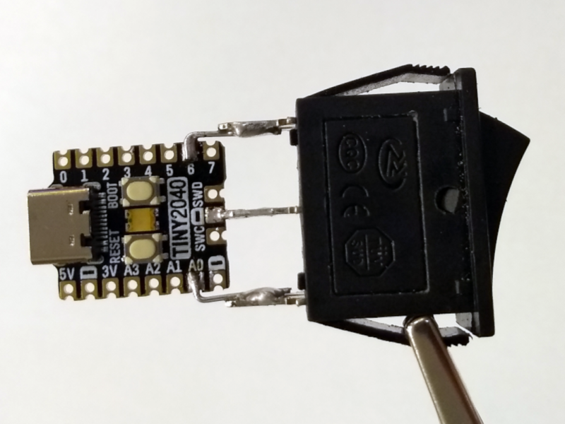The TinyPICO board and the rocker switch soldered together showing a complete device, shown being held in the air by a crocodile clip