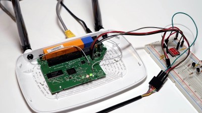 A TP-Link router with its cover taken off, jumper wires going from its PCB to a breadboard, that a logic level shifter PCB is plugged in. Other set of wires from the shifter then seems to go into a USB-UART adapter.