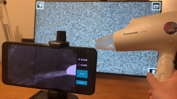 On the left side, there's a smartphone. On the right side, there's a hairdryer turned on. On the smartphone screen, you can see the working end of the hairdryer shown, as well as a jet of air coming out of that end. In the background, there's an LCD screen showing a noise pattern.