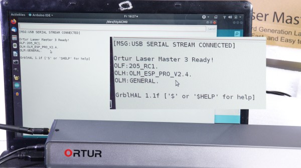 Showing an Ortur lasercutter control module in front of a screen. There's a serial terminal open on the screen, showing the "Ortur Laser Master 3" banner, and then a Grbl prompt.