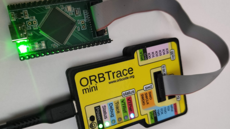 The Orbtrace debugger hardware connected to a development board t hrough a 20-pin ribbon cable. The development board has a green LED shining.