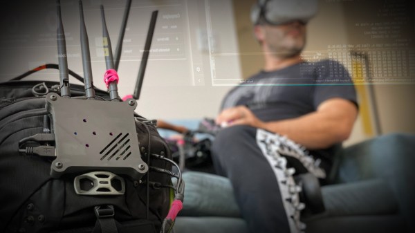A person sits on a couch in the background wearing a VR headset. A keyboard is on their lap and a backpack studded with antennas and cables sits in the foreground.