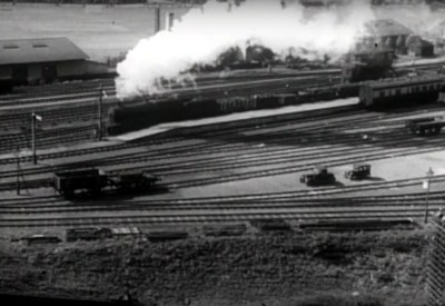 A steam train passing through a station, from a distance in black and white