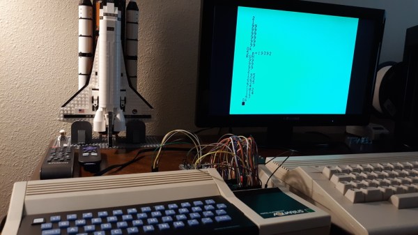 A small keyboard form factor retrocomputer with blue keys on a black background sits in front of a display and a LEGO model of the Space Shuttle. There are a number of jumper wires and a breadboard coming from an open panel on the right side of the machine.