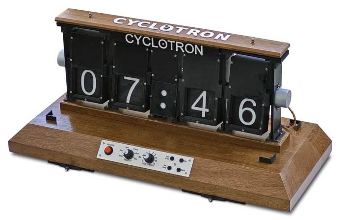 A cyclotron clock display, mounted on a wooden base. There are two stepper motors exposed on either side. There is a panel installed in the wooden base with a red button on the left, two knobs and four smaller buttons in a two by two grid on the right.