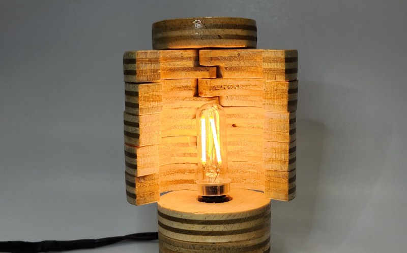 A segmented lamp made of circular slices of plywood. They are arranged as shutters around a long, skinny LED bulb in the center that gives off an incadescent-looking glow. A cord trails off to the left against the grey background.