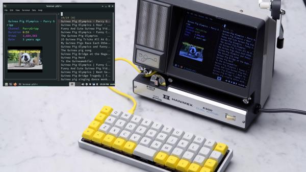 A terminal window with a search for "Guineau Pig Olympics" is inset on a photo of an ortholinear keyboard attached by a yellow USB cable to a 70s aluminum and plastic Super 8 film editor/viewer. The device has a large screen on the right hand side, a silver grate on the left, and a tray at the bottom for slotting in film.