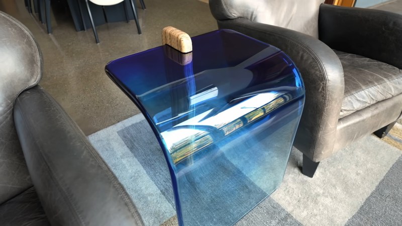 A transparent blue resin waterfall tabletop with a single wooden leg sits on a grey rug between two grey leather chairs.
