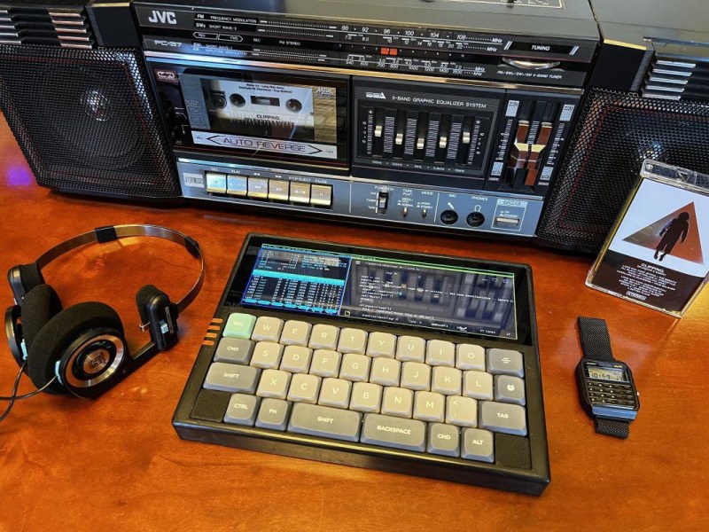 A widescreen slate-style cyberdeck with a small keyboard sits in front of a cassette deck stereo. Headphones sit to the left of the deck and an old Casio calculator watch is to the right.