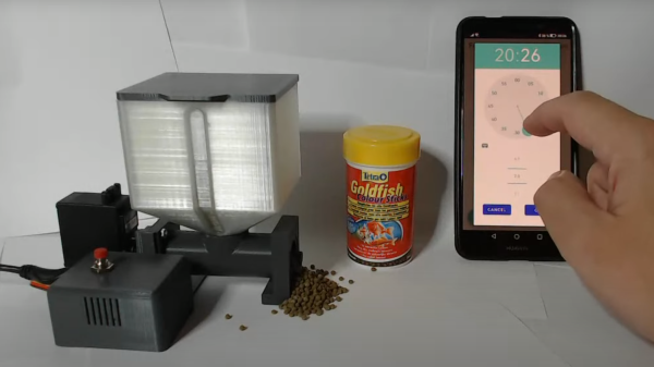 3d printed fish feeder system with food basin, electronic housing with red button on top and servo attached on the side. A pile of food is coming out of the 3D printed fish feeder mechanism. In the middle of the picture is a can of goldfish pellet food. On the right is a hand interacting with a propped up cell phone, setting a time.