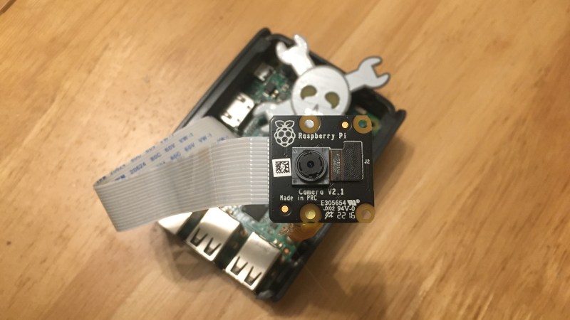 A Raspberry Pi 3 with a black Raspberry Pi Camera PCB on top of it, looking at the camera taking this picture. There's a Jolly Wrencher in the background.