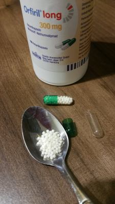 An Orfiril medication bottle is shown, with an Orfiril pill capsule next to it, showing the small pellets inside. Another pill capsule has been disassembled, with the pellets inside a teaspoon.