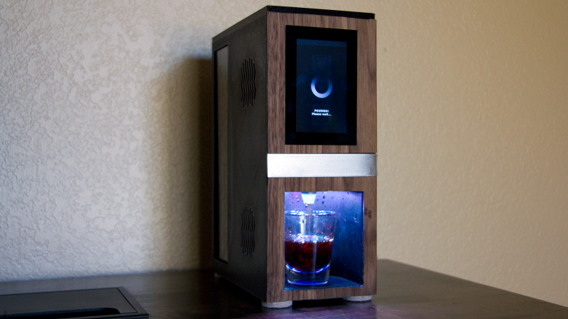 A picture of the JagerMachine consisting of rectangular, desktop sized drink serving machine with a wooden varnish, a 3.5 inch touchscreen on the front face on top and a cavity with a shot glass in it, lit up by blue leds, with liquid pouring into it.