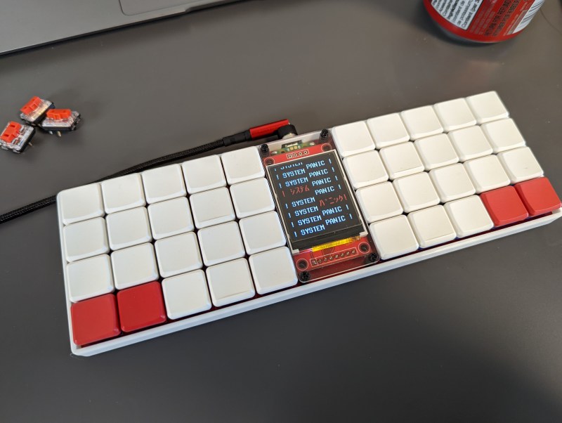 An ortholinear keyboard with predominantly blank white keycaps. There are two red keycaps on the bottom outside corners. The center of the keyboard houses a large LCD in portrait orientation on a red PCB.