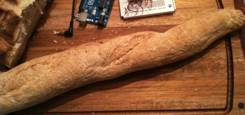 A baguette sits diagonally across a wooden cutting board. Above it sits an Arduino and an electronics breadboard.