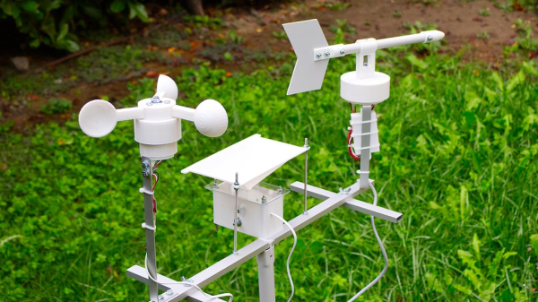 An esp32 weather station with 3d printed anemometer, rain gauge and wind vane mounted on an aluminum frame sitting in an overgrown lawn