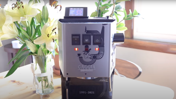 A Gaggia classic espresso machine with an LCD screen attached to the top, sitting on a table with a vase of yellow lily flowers on the left and sunlight coming in from a window on the right.