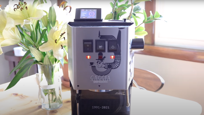 A Gaggia classic espresso machine with an LCD screen attached to the top, sitting on a table with vase of yellow lily flowers to its left and sunlight coming in from a window from the right.