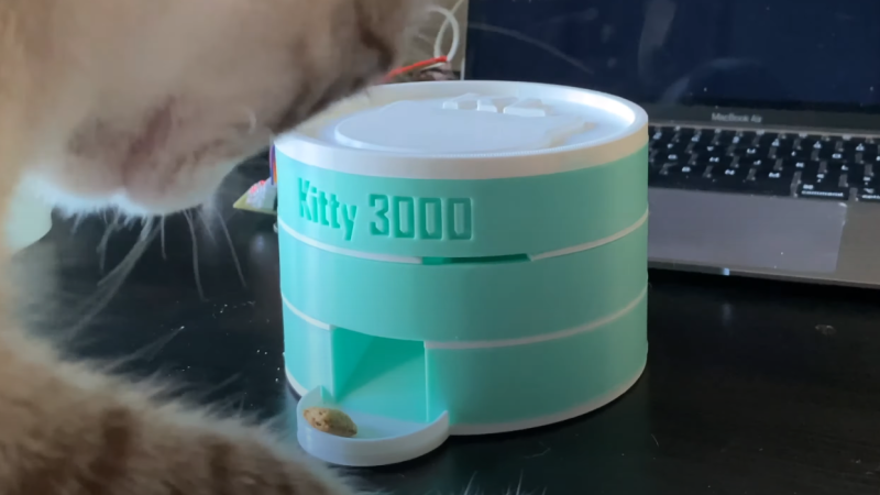 A 3D printed cat treat dispenser on a table with a laptop in the background and with a treat in it's tray and a cat on the left about to eat the treat.