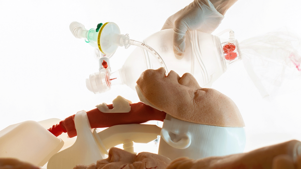 A profile view of a medical training mannequin with a tube down its "throat." A ventillation bag is in the gloved hand of a human trainee.