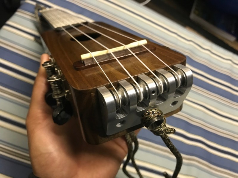 A walnut ukulele with an aluminum piece routing strings at it's base which is facing the camera. The neck of the instrument extends away from the viewer and is held at an angle by a hand with striped sheets in the background.