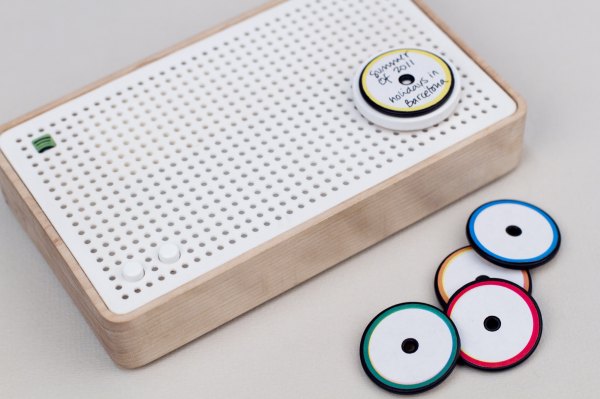 A radio with a white front grate and wood edges sits on a grey surface. Next to the radio are small white disks with colorful edges reminicient of microdisc-sized records. A yellow-ringed disk sits on the radio. The handwritten title says, "Summer of 2011; Holidays in Barcelona"