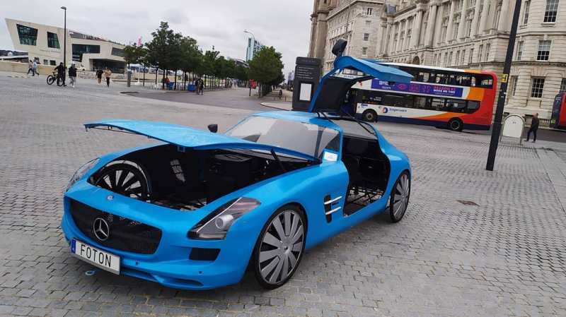 A blue Mercedes SLS AMG sports car body with bicycle wheels. The gull wing is open to reveal the spartan interior and the hood is open to reveal an empty engine compartment since this is actually a bike.