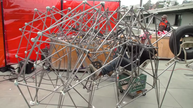 TV personality and maker, Adam Savage, sits on a chair attached to a milk crate on wheels. It is situated inside an assortment of steel tubes forming the legs and body of a strandbeest walking machine.