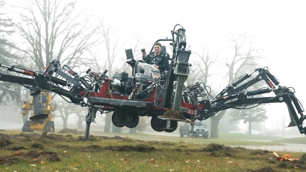 A man sits in a chair atop a hexagonal platform. From the platform there are six hydraulically-actuated legs supporting the hexapod above a grassy field. The field is filled with fog, giving the shot a mysterious, otherworldly look.