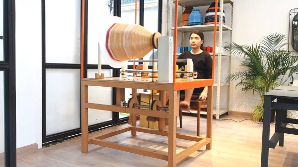 A woman sits at a wooden table with a set of pedals attached. A large frame sits on top of the table with a lampshade form spinning in it and five strings run through an apparatus to the frame. A shelving unit with finished lampshades sits behind the woman.