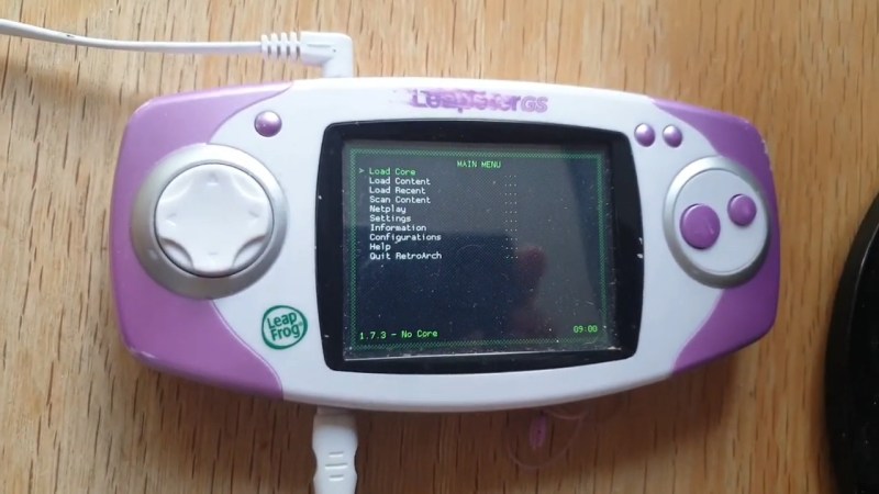 A pink and white Leapster GS handheld console sits on a wooden table. It has a white D-pad and two large pink action buttons. A power cord extends from the bottom and a headphone cable comes out the top.