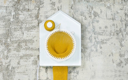 A white clock with a house profile sits on a variegated grey background. A yellow skein of yarn sits on the top left side of the clock feeding into a circular loom that takes up the bulk of the center. A yellow scarf extends out the back of the clock and out of frame below the image.