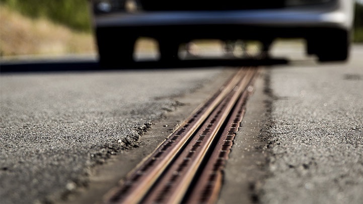 A grey car sits in the background out of focus, its front facing the camera. It sits over an asphalt roadway with a metal rail extending from the foreground to behind the car in the distance. The rail has a two parallel slots and screws surrounding the slots running down the rail.