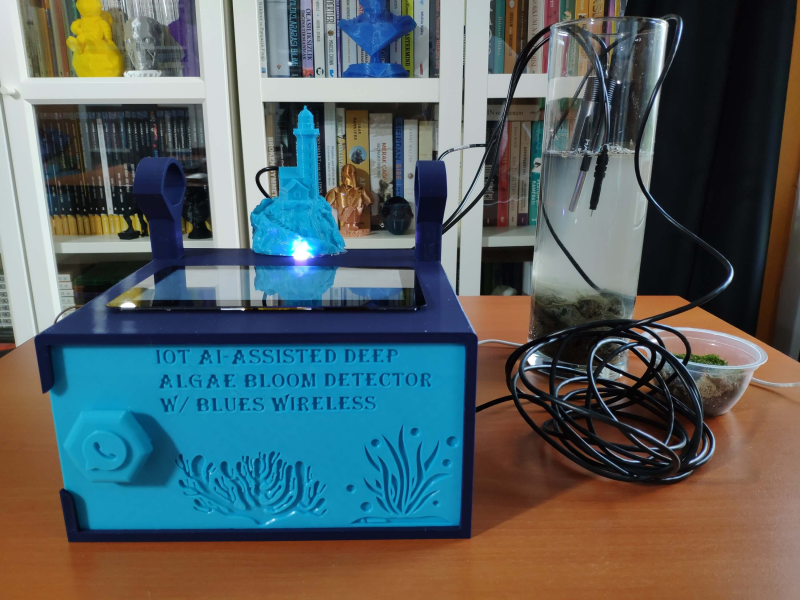 A blue enclosure with "IoT AI-assisted Deep Algae Bloom Detector w/Blues Wireless" written on the front. Two black cables run over a wooden desk to a cylinder with rocks on the bottom and filled with murky water. A bookshelf lurks in the background.