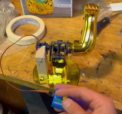 A golden maneki-neko cat arm mechanism attached to a servo on a workbench with a hand controlling a servo motor tester that's plugged into the servo attached to the arm.