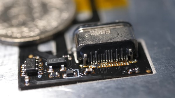 A closeup of a black flexible PCB with an out-of-focus quarter in the background, approximately the same size as the end of the PCB we're looking at. One the right is a USB C connector and to its left are two SMD components with visible pins. Several smaller SMD components (resistors or caps?) are soldered to other parts of the board.