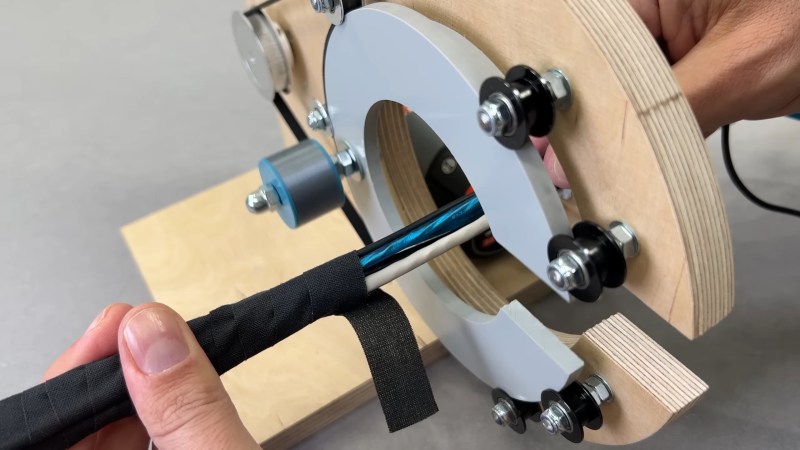 A person holds a bundle of white, black, and blue wires. The left hand side of the wires are wrapped with black tape. The wires are inside a wire wrapping machine with a grey plastic "C" which rotates inside seven small pulleys. A large pulley in the background drives three of the pulleys to rotate the "C" around and wrap the wires with tape from the spool attached to the "C."