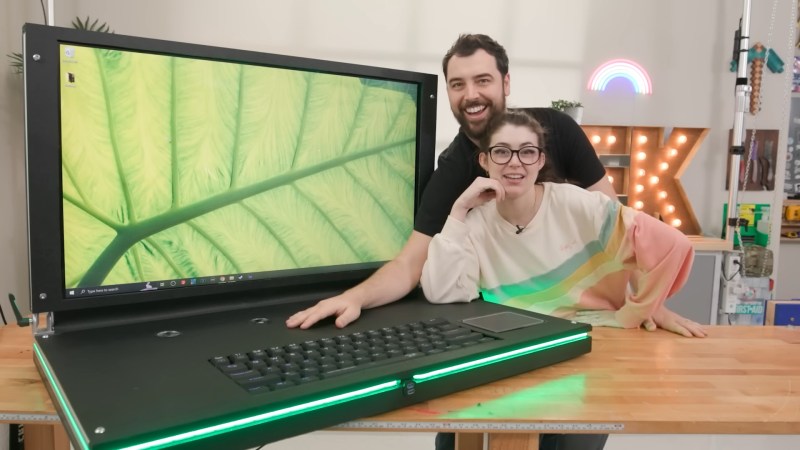 Two people lounge over a wooden tabletop to lean on a large black laptop. It has a green leaf on its 43" LCD desktop and RGB lighting around its edge is glowing a slightly deeper shade of green.
