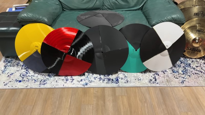 A series of five cymbals sitting on white and black speckled carpet in front of a green loveseat. Each cymbal is assembled from four printed sections. Their colors from left to right are yellow and grey, red and black, black, teal and black, and white and black. A sixth, grey and black cymbal is sitting in the middle of the loveseat cushion.
