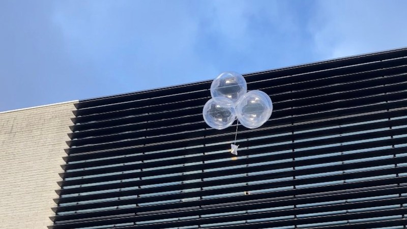 Showing balloon rising up, not too far from the ground, with one of the FOSDEM buildings and sky in the background