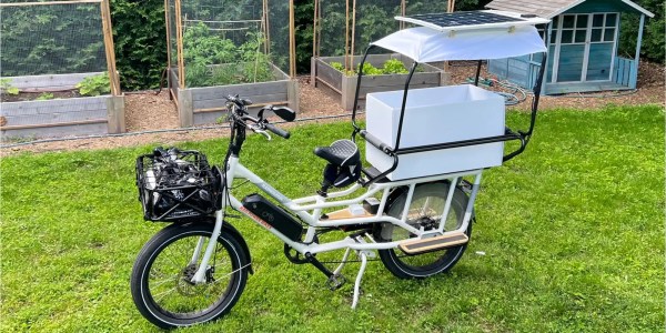A white longtail cargo bike sits on grass with fenced-in planters behind it. The bike has a basket made of black metal tubes on the front and a passenger compartment behind the rider seat for children made of similar black metal tubes. A white canopy is above the passenger compartment and a solar panel sits atop the canopy.