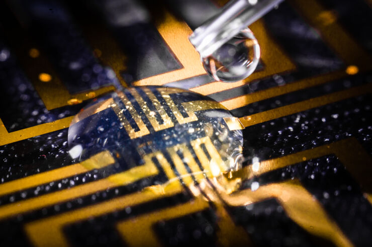 A clear droplet sits on a blue PCB with gold traces. A syringe with a drop of clear liquid sits above the droplet.