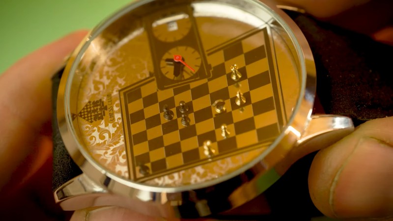 A metal watch is held in a man's fingers. The watchface has a laser etched chess board with miniature chess pieces made of brass enacting a match. The time is told on an etched chess clock to the right hand side of the timepiece and a small window on the rightmost "clock" shows the date.