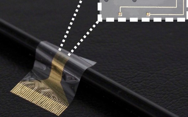 A clear flexible PCB with a number of gold electrodes on one end. It is wrapped over a black cable to demonstrate its flexibility. A set of dashed white lines goes from one end to a zoomed in image of the circuit structure inset in the top right of the image.