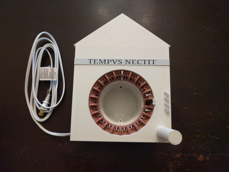 A white, house-shaped clock with the words "TEMPUS NECTIT" written in faux Roman script in black on a strip of silver at the base of the "roof." a white power cord extends from the left of the enclosure, and the center of the clock is a 22 pin knitting machine wheel with one pin covered in silver metalic. A white plastic peg extends from the bottom right of the enclosure to hold the feedstock yarn.